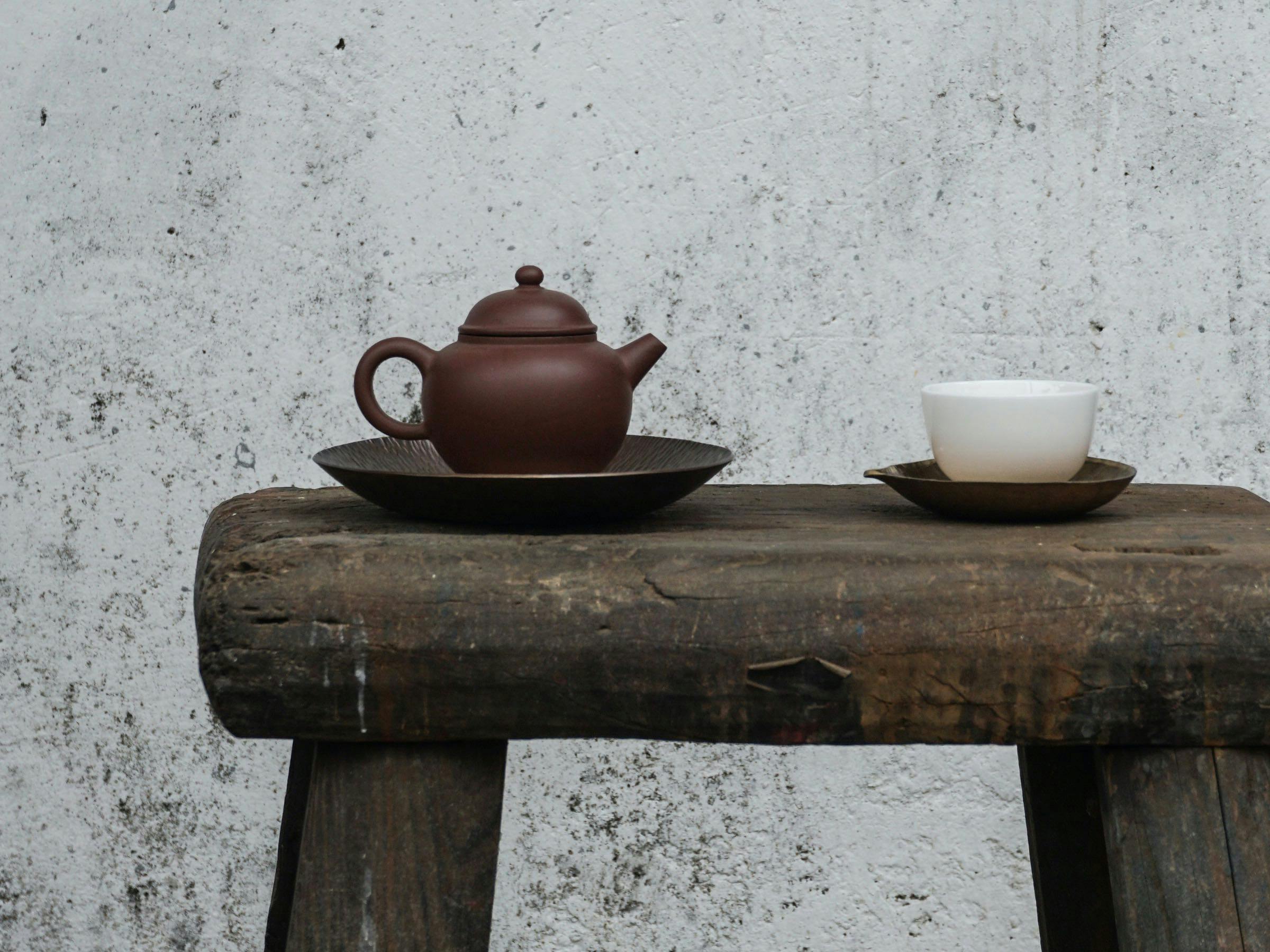 Teekanne aus Ton und Tasse aus Keramik auf einem Hocker aus Holz vor einer grauen Wand